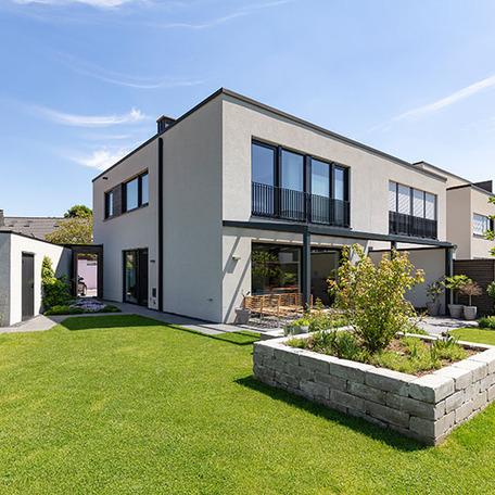 Wohnungen mit weisser Fassade aus der Perspektive im angelegten Garten mit Terrasse mit Dach und Moebeln, Garage, Auffahrt mit Auto