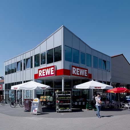 Eingang des Fachmarktzentrum in Recklinghausen von aussen mit grauer Fassade, Rewe Logo, einigen zuverkaufenden Produkten in Regalen und Sonnenschirmen
