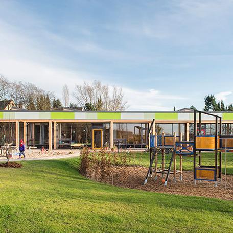 Spielplatz mit Klettergeruest, Sandkasten und dem gruenen Gebaeude der Kindertagesstaette im Hintergrund