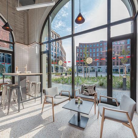 Foto von einem Raum mit Moebeln welches einem Cafe gleicht und durch die Fensterfront die angelegten Beete und angrenzenden Gebaeude des Gewerbecampus Ovum in Koeln mit einfaengt