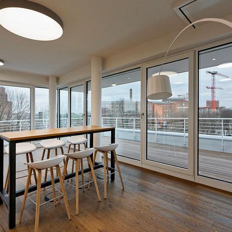Raum mit Holzboden, Holzstehtisch und sechs Hockern in der Naehe einer Fensterfront durch die man auf den Balkon treten kann des Erweiterungsbau eines Reiseveranstalters