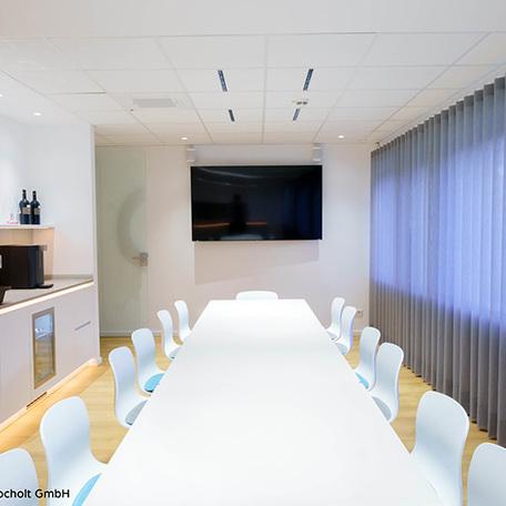 Meetingraum mit langem weissem Tisch, Stuehlen und einem grossem Fernseher an der weißen Wand der sanierten Gewerbeflaeche in Bocholt