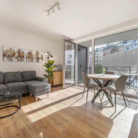 Wohnzimmer einer moeblierten Wohnung in Duesseldorf Lindenstrasse mit grauer Couch mit runden Couchtisch aus Glas, runden Esstisch mit Stuehlen, Dekoration und offender Glastuer die auf den Balkonfuehrt