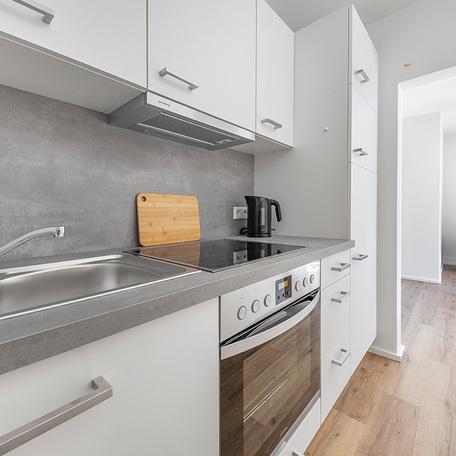 Kueche einer moeblierten Wohnung in Duesseldorf Lindenstrasse mit weissen Fronten, Kuehlschrank, Ofen, Spuelbecken, Herd, Dunstabzugshaube, viel Stauraum in Schraenken, Wasserkocher und Holzbrett