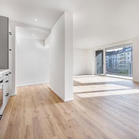 Wohnzimmer mit offener grauer Kueche einer Wohnung in Duesseldorf Lindenstrasse mit weissen Waenden, Boden in Holzoptik, Fenster und Glastuer welche auf den Balkon fuehrt