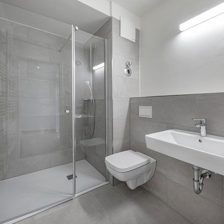 Badezimmer einer Wohnung in Duesseldorf Lindenstrasse mit grauen Fliesen, Waschbecken, Toilette, Beleuchtung und Dusche