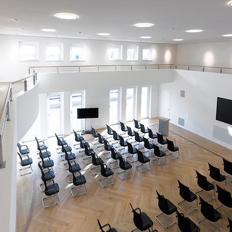 Blick aus der zweiten Etage auf den Versammlungsraum des Verwaltungsgebaeude in Duisburg mit Holzboden, schwarzen Stuehlen, Rednerpult, Fernseher und weissen Waenden