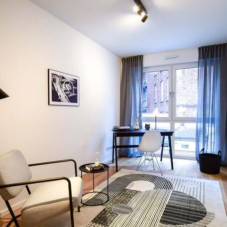 Arbeitszimmer einer Musterwohnung des Bauprojektes Duesseldorf Oberbilk mit Arbeitstisch, Arbeitsstuhl, Sessel mit Tischchen, schwarzen Vorhaengen, Tippich und Bild an der weiss tapezierten Wand