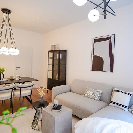 Wohnzimmer einer Musterwohnung des Bauprojektes Duesseldorf Oberbilk mit grauem Sofa, Couchtischen, Bild an der weiss tapezierten Wand, schwarzer Vitrine und Esstisch im Hintergrund