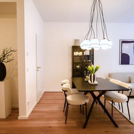Wohnzimmer einer Musterwohnung des Bauprojektes Duesseldorf Oberbilk mit schwarzem Esstisch, beigen Stuehlen, schwarze Vitrine, aesthetischer Lampe ueber dem Tisch, weisser Tapete und Boden in Holzoptik