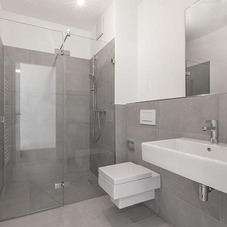Badezimmer einer Wohnung des Wohnquatiers in Angermund mit grauen Fliesen, weisser und Edelstahlausstattung, Dusche, Toilette, Waschbaecken und Spiegel