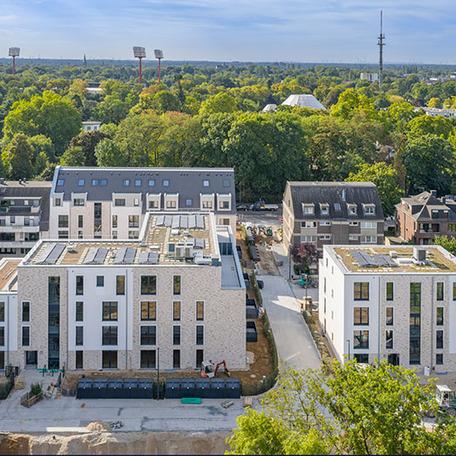 Drohnenansicht der drei Wohngebaeude mit beiger Fassade, Solaranlagen auf dem Dach und Einfahrt zum Parkplatz im Hinterhof