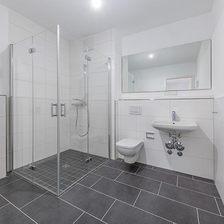 Badezimmer einer Wohnung in Krefeld mit weissen Fliesen and den Waenden, grauen Fliesen auf dem Boden, Dusche, Toilette, Waschbecken und Spiegel