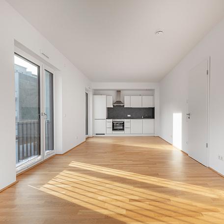 Wohnzimmer mit weisser Kueche einer Wohnung des Wohnquatiers in Moers mit weissen Waenden, Holzboden, Fensterfront mit Tuer und Balkon