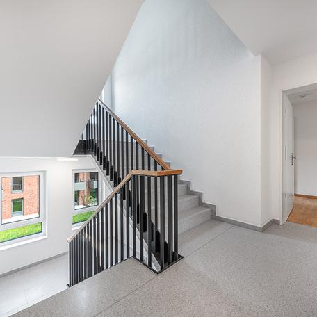 Treppenhaus eines Gebaeudes des Wohnquatiers York in Muenster mit grauem Boden, weissen Waenden, grauem Gelaender, zwei Fenstern und eine Tuer einer Wohnung offen